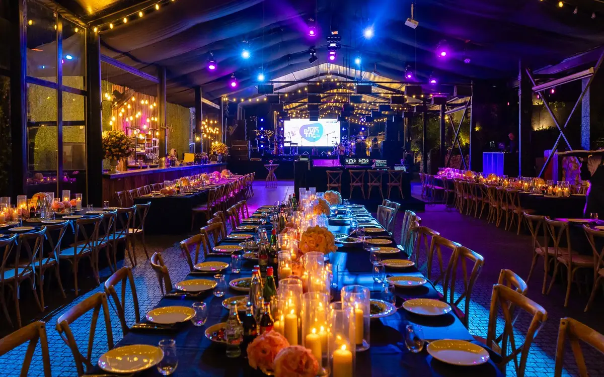 Long candlelit dinner tables in an event venue lit in blue and violet, a stage screen at the far end