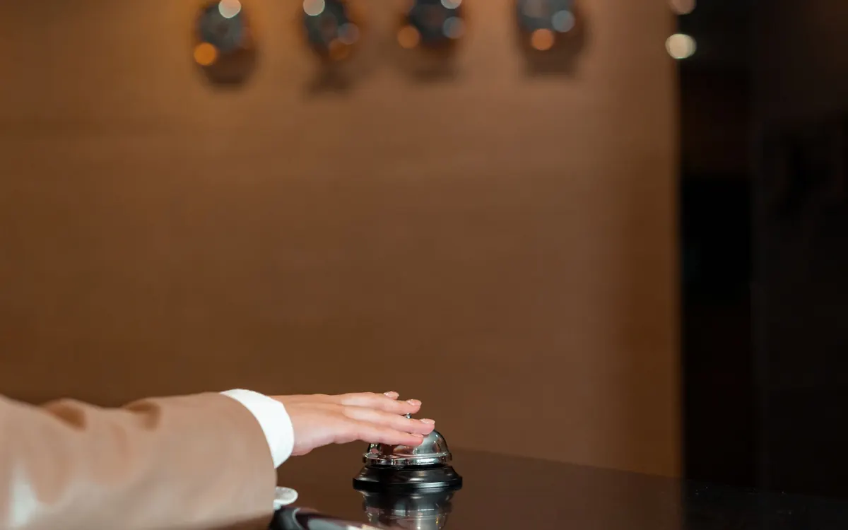 A hand ringing a silver service bell on a dark reception desk, warm lights blurred behind
